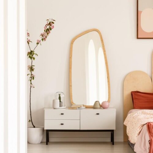 Plant next to white dressing table with mirror in red bedroom apartment interior with poster above bed.