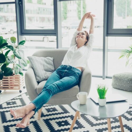 Weekends finally! Relaxed sleepy serene lady is sitting on modern chair near the big windows in light cozy room at home. She is enjoying peace and morning, stretching her arms and foot