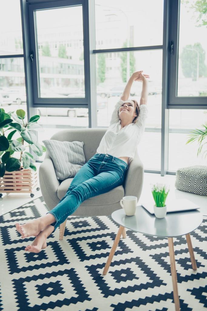 Weekends finally! Relaxed sleepy serene lady is sitting on modern chair near the big windows in light cozy room at home. She is enjoying peace and morning, stretching her arms and foot